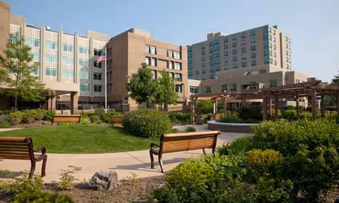 Outdoor courtyard with benches, pathways, and landscaped greenery outside a large care facility.
