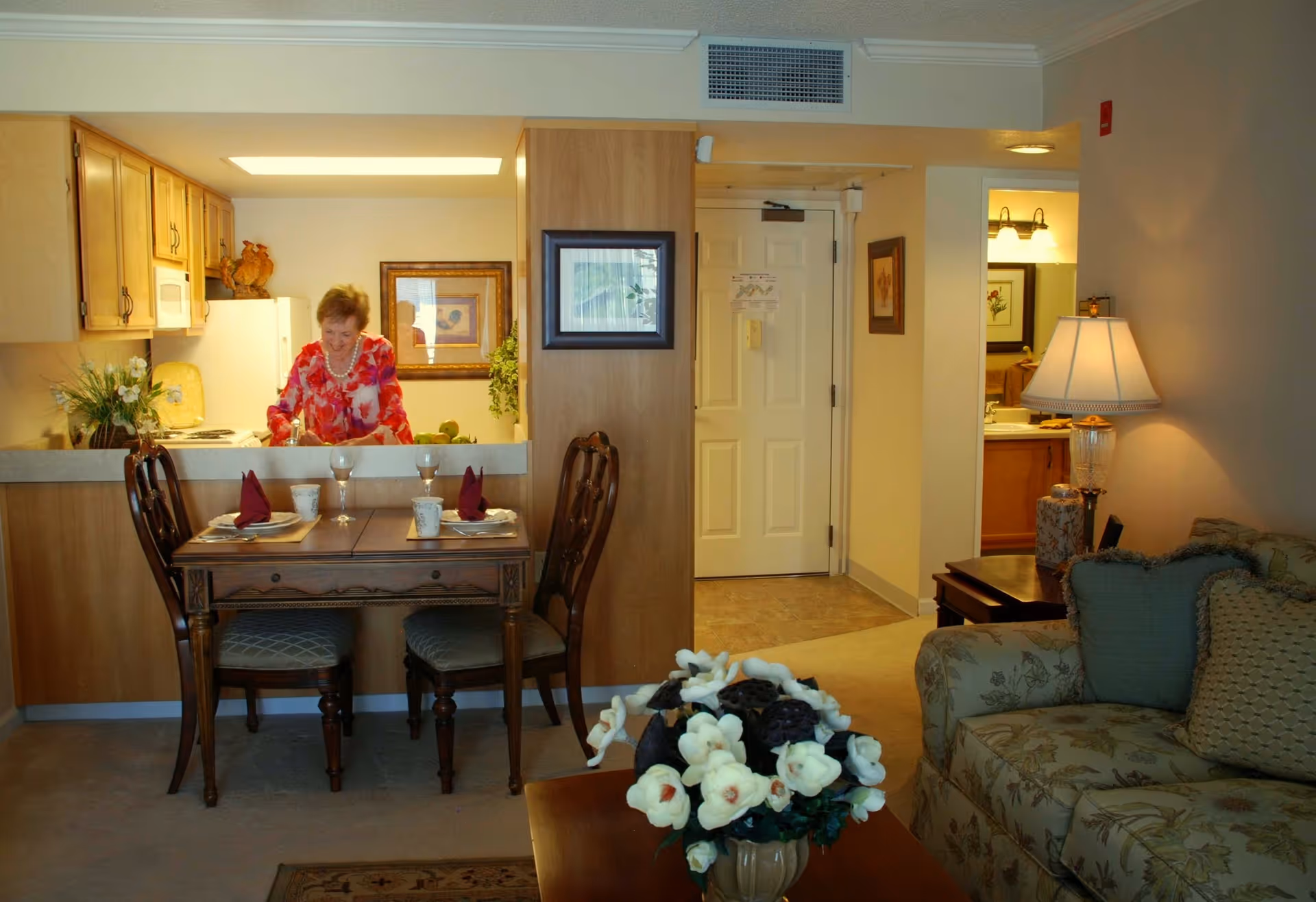Interior view of a senior living facility apartment showing a woman in a red floral blouse standing in a kitchen area behind a counter. In front of the counter is a small dining table set for two with folded napkins and cups. To the right is a living room area with a floral-patterned sofa, a side table with a lamp, and a coffee table with a vase of white flowers. The entrance door and a bathroom doorway are visible in the background.