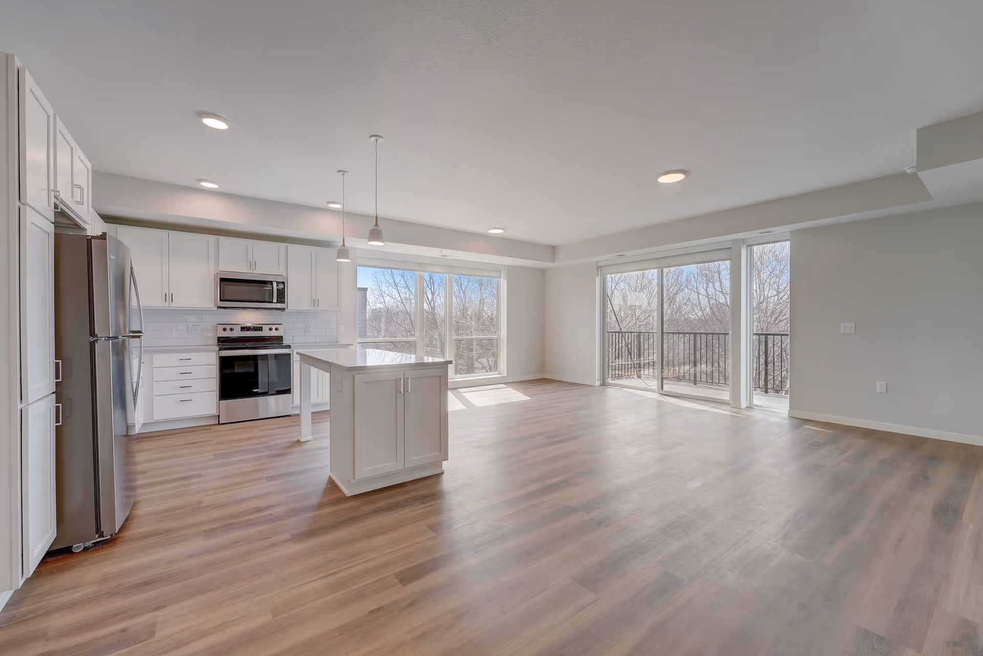 Bright and spacious open-concept kitchen and living area with large windows and sliding glass doors leading to a balcony. The kitchen features white cabinetry, stainless steel appliances, a central island, and wood flooring throughout.