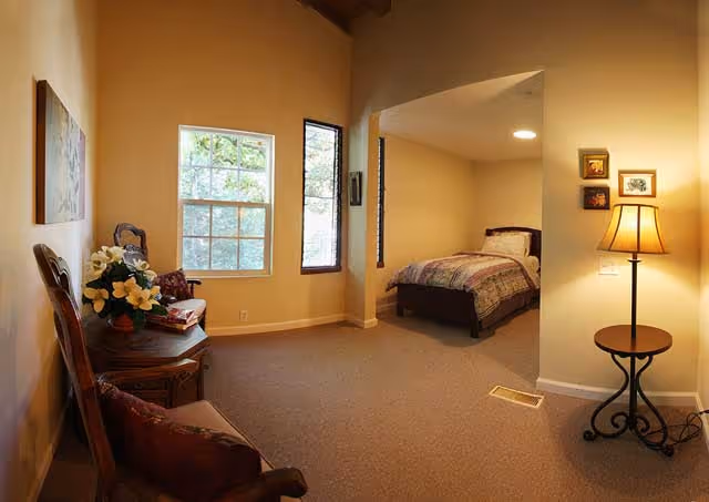 Bright bedroom with a bed in an alcove, a seating area with chairs and a side table topped with flowers, and a floor lamp by framed pictures.