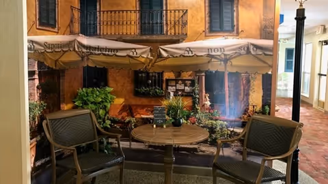 Indoor seating area with two brown chairs and a round table in front of a wall mural depicting an outdoor café scene with umbrellas, plants, and a building facade.