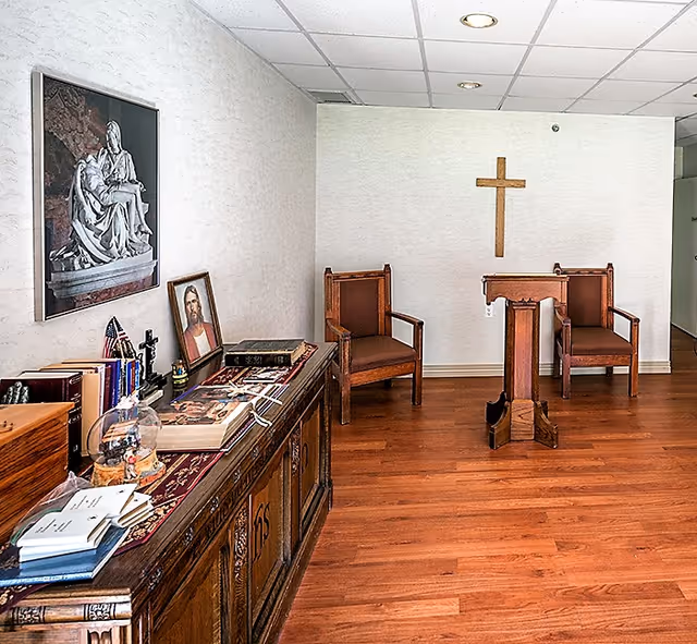 A small chapel or prayer room with wooden flooring, two wooden chairs, a wooden lectern, and a wooden cross mounted on the wall. A sideboard holds religious books, a framed picture of Jesus, a small American flag, and other religious items. A framed picture of a religious sculpture hangs on the wall.