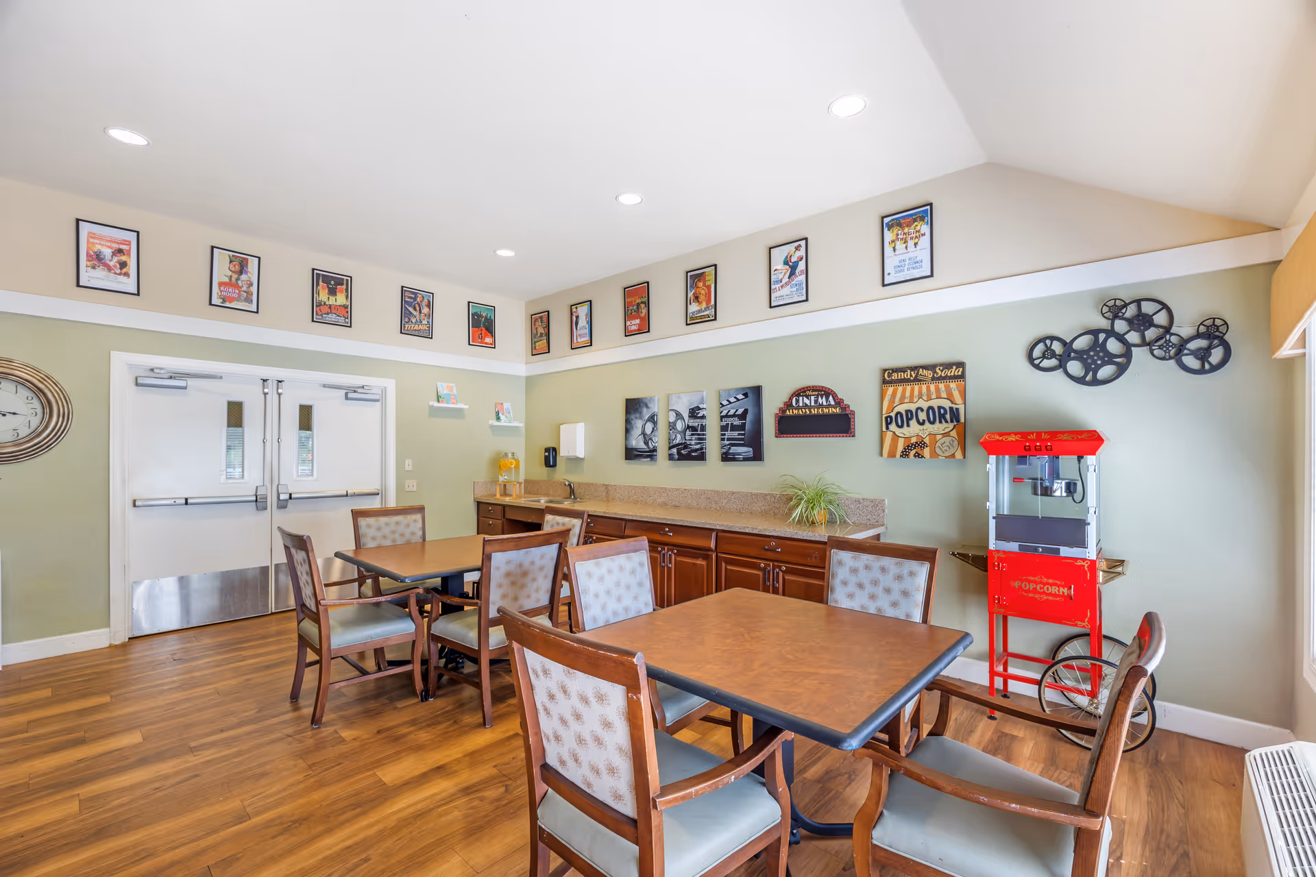 A cozy dining area with wooden floors and light green walls. There are two tables with six cushioned chairs arranged around them. On the right side, there is a red popcorn machine and wall decorations including movie-themed signs and film reel art. Above the double doors on the left wall, there are framed vintage movie posters. The room has recessed lighting in the ceiling and a countertop with cabinets underneath along the back wall.