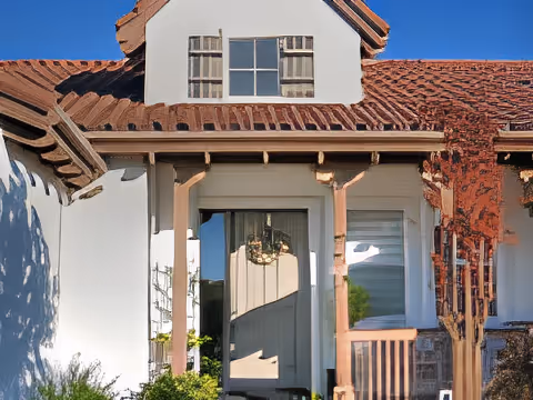 Front entrance of a stucco house with a tile roof, wooden porch posts, a wreath on the door, and a small shuttered window above.
