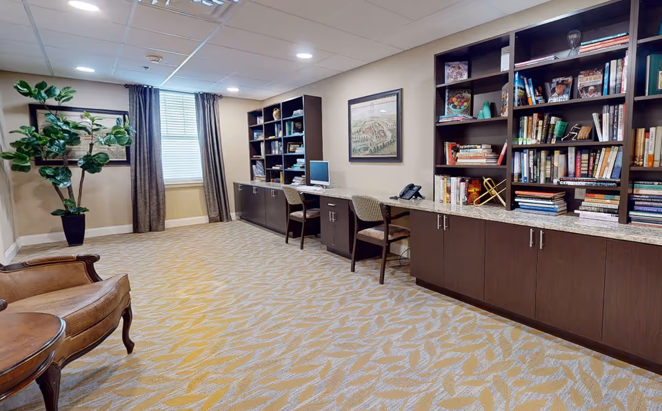 A quiet reading and work area in a senior living facility featuring built-in dark wood bookshelves filled with books and decorative items. There are two chairs positioned at a long granite countertop with a computer and a phone. The room has beige walls, a window with curtains, patterned carpet, and a potted plant in the corner.
