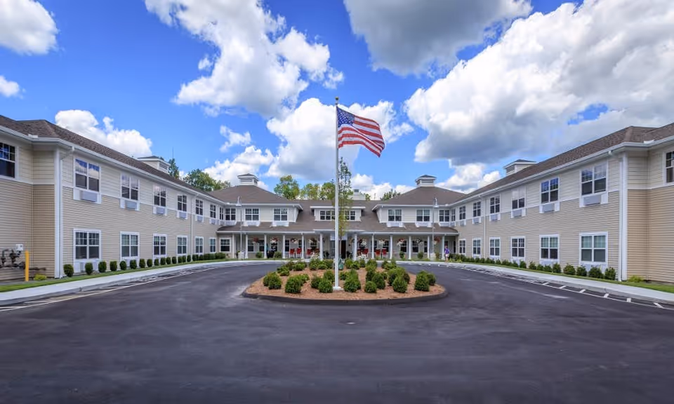 Front exterior view of a two-story assisted living facility building with beige siding and white trim. There is a circular driveway with a landscaped island in the center featuring shrubs and an American flag on a flagpole. The sky is blue with scattered clouds.