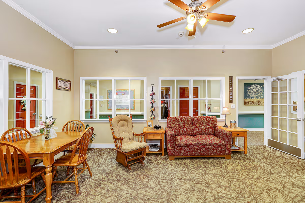 A cozy common area in a senior living facility featuring a wooden dining table with four chairs, a cushioned rocking chair, a red floral-patterned loveseat, two wooden side tables with lamps, and a ceiling fan with lights. The walls are beige with white trim, and there are large windows and glass doors providing views into adjacent rooms.