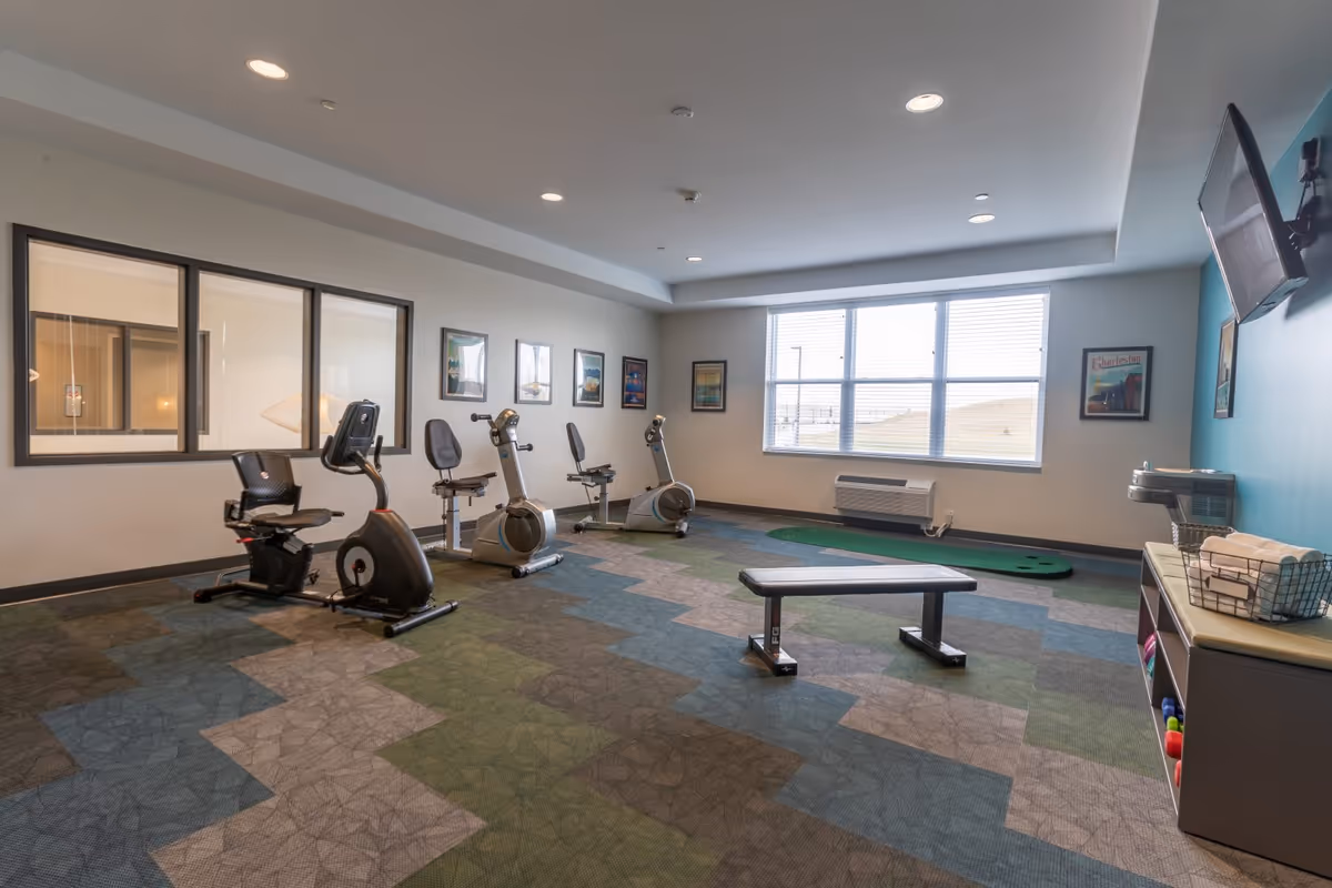 A fitness room with exercise equipment including three stationary bikes, a workout bench, and a putting green near a large window. The room has patterned carpet in shades of blue, green, and gray, framed pictures on the walls, a wall-mounted TV, and a shelf with towels and small exercise weights.