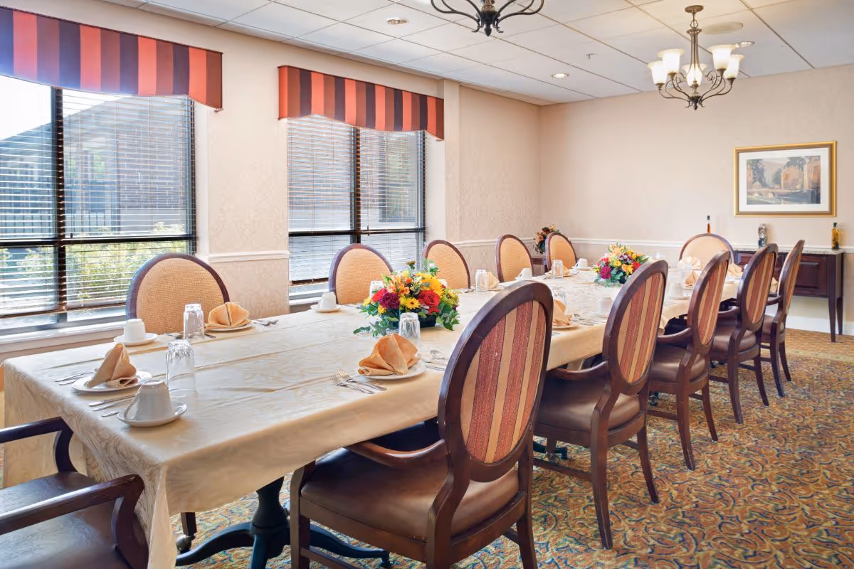 A formal dining room with a long rectangular table covered with a beige tablecloth. The table is set with folded napkins, cups, glasses, and silverware. There are floral centerpieces placed along the table. The room has large windows with striped valances, patterned carpet, and a chandelier hanging from the ceiling. A framed picture hangs on the wall in the background.