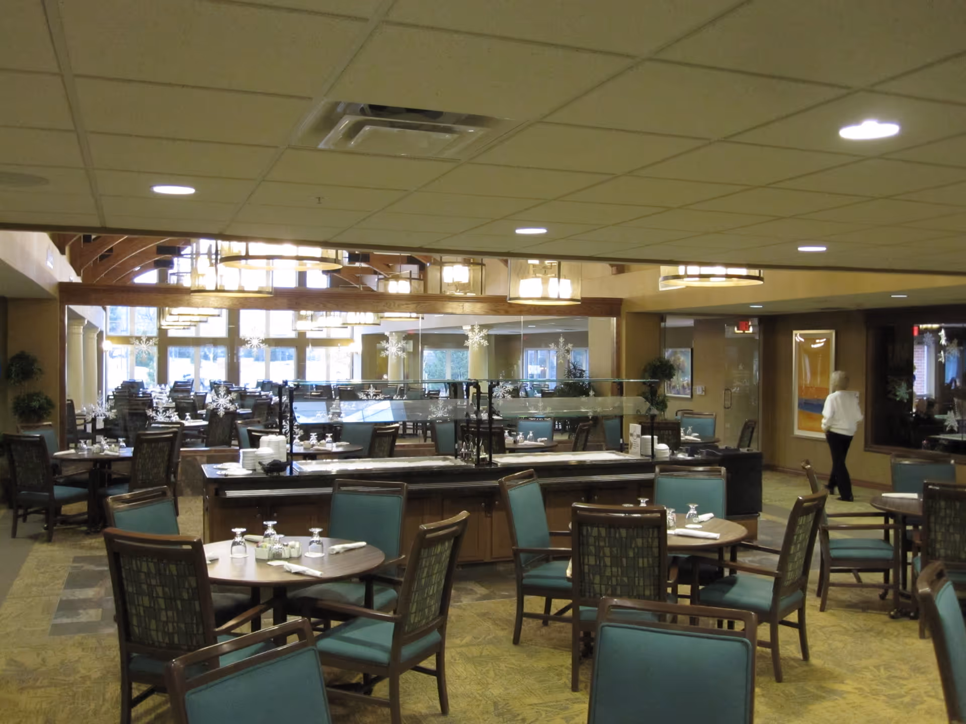 Interior view of a dining room in a retirement community with multiple round tables and chairs arranged neatly. Tables are set with glasses, napkins, and condiments. The room features large windows, decorative lighting fixtures, and a central buffet area with glass sneeze guards. A person is walking in the background near the entrance.