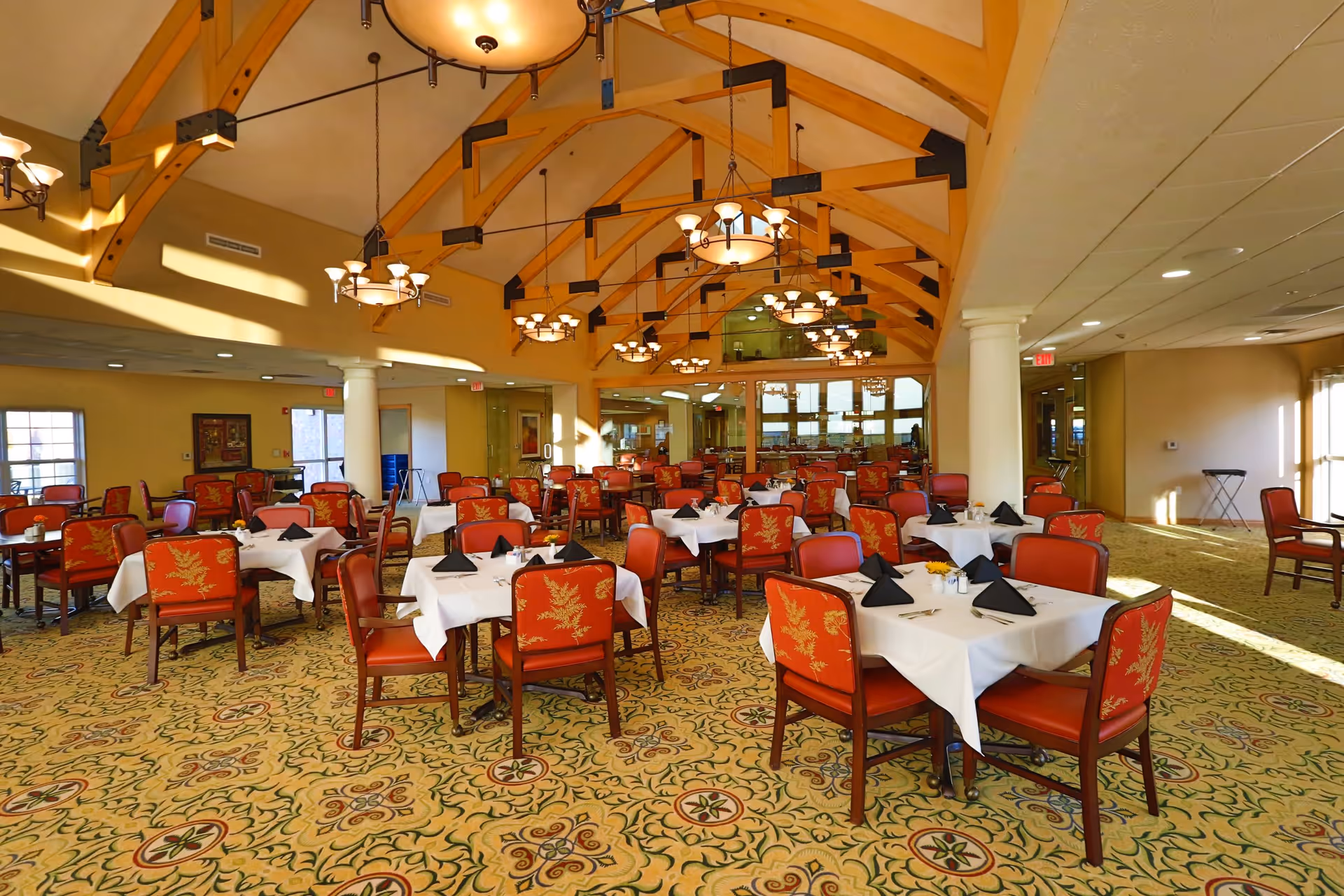 A spacious dining room with multiple tables covered in white tablecloths and set with black folded napkins. The chairs have red upholstery with a gold leaf pattern. The room features a high ceiling with exposed wooden beams and several hanging chandeliers providing warm lighting. Large windows allow natural light to fill the space, and the carpet has an intricate floral design.