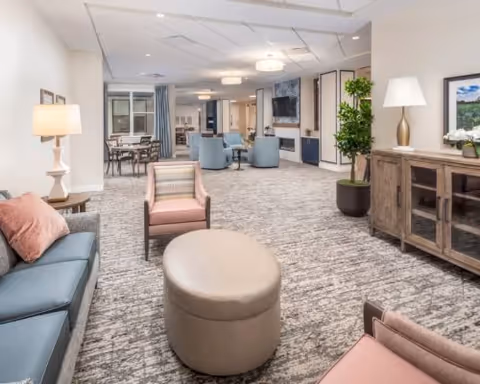 Spacious, well-lit senior living lounge with sofas, armchairs, an ottoman, a TV, sideboard and dining tables in the background.