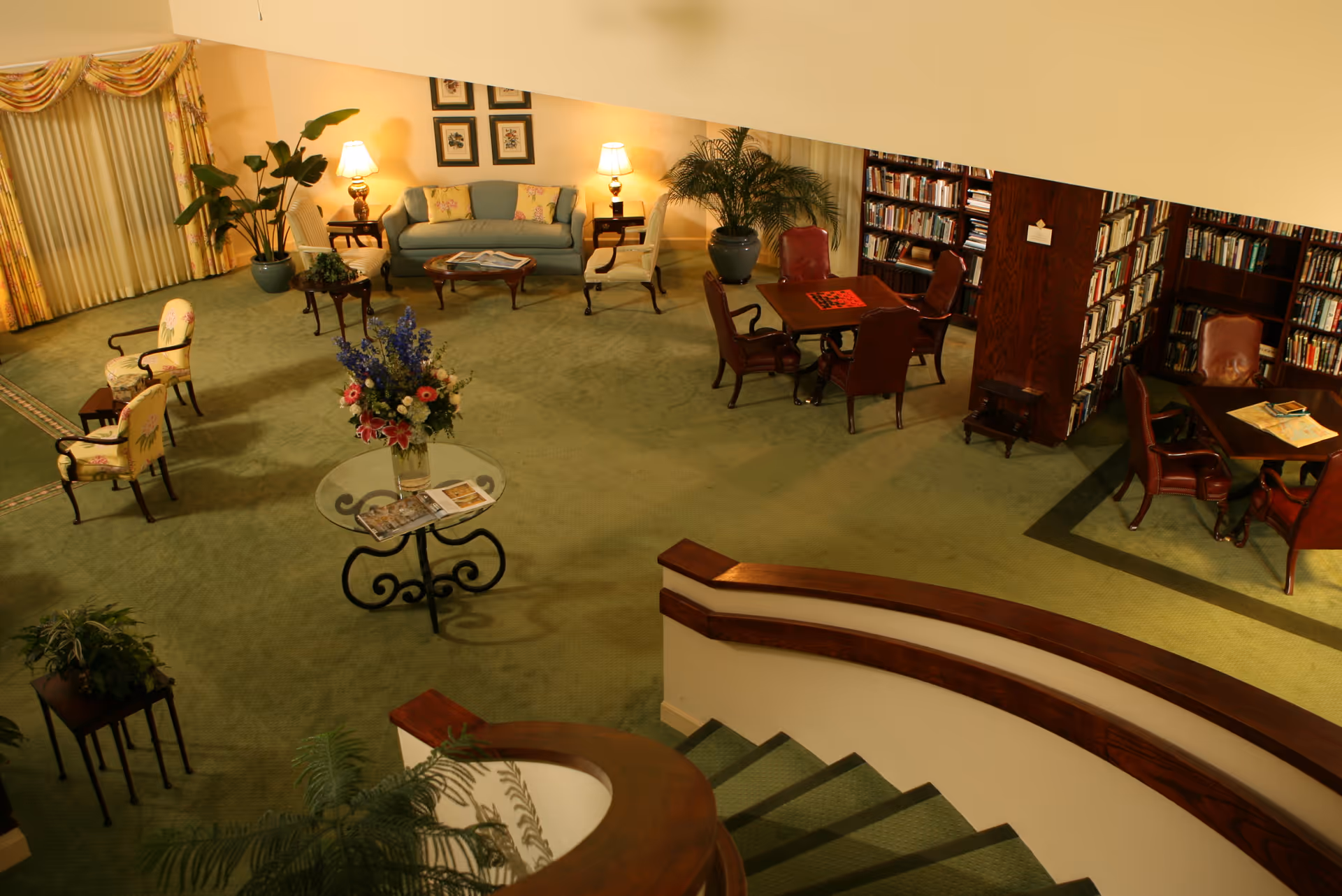 Interior view of a cozy common area with green carpet, a curved staircase with wooden railing, a glass table with a flower arrangement, several upholstered chairs, a sofa with side tables and lamps, and bookshelves filled with books. There are also tables with chairs for reading or playing games.