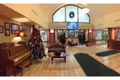 A spacious lobby area decorated for Christmas with a large Christmas tree, a piano, a coffee table with a centerpiece, and a television mounted on the wall above a fireplace. The room has tiled floors, green rugs, framed pictures on the walls, and large windows letting in natural light.