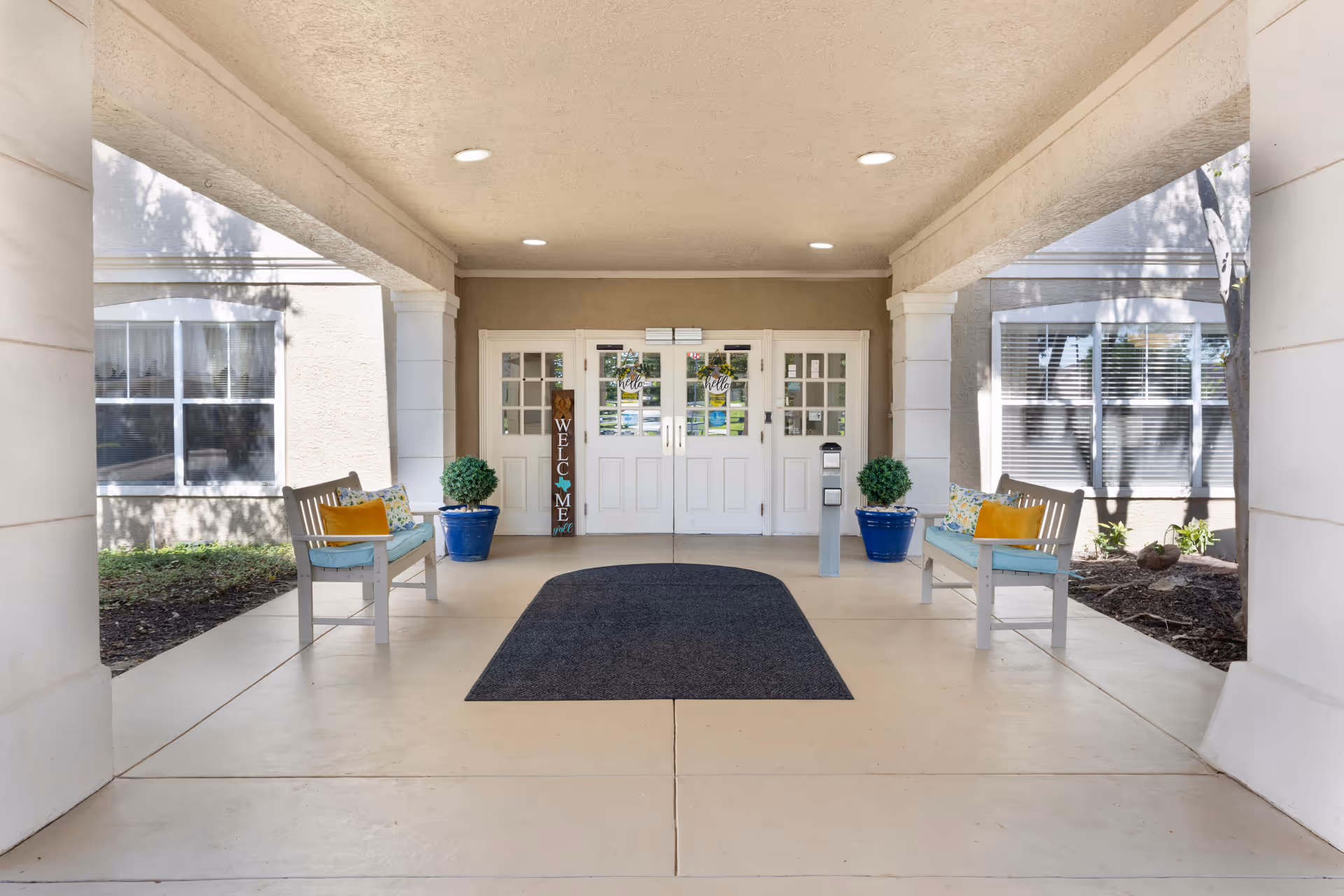 Covered entrance area of a building with double white doors decorated with welcome signs. Two benches with cushions and pillows are placed on either side, along with blue potted plants. The area is well-lit with recessed ceiling lights and has a dark floor mat in front of the doors.