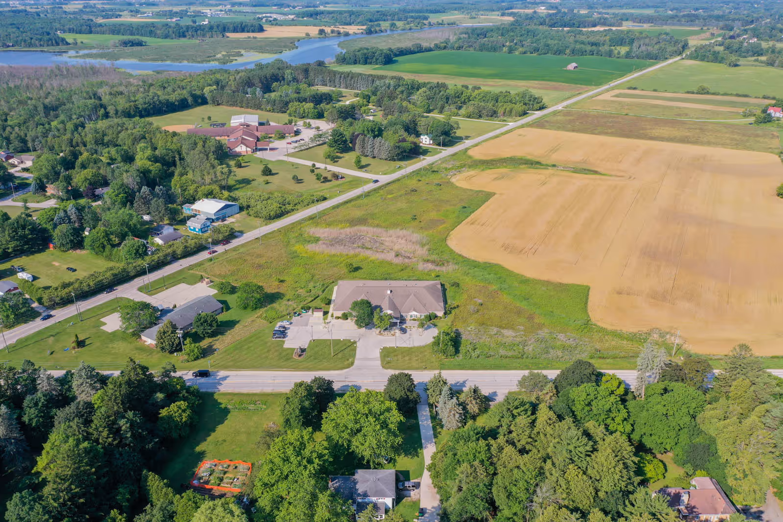 Aerial view of Meadow View Assisted Living facility surrounded by green trees, open fields, and a river in the distance. The building is located near a road with parking spaces and is set in a rural landscape with other buildings and farmland nearby.