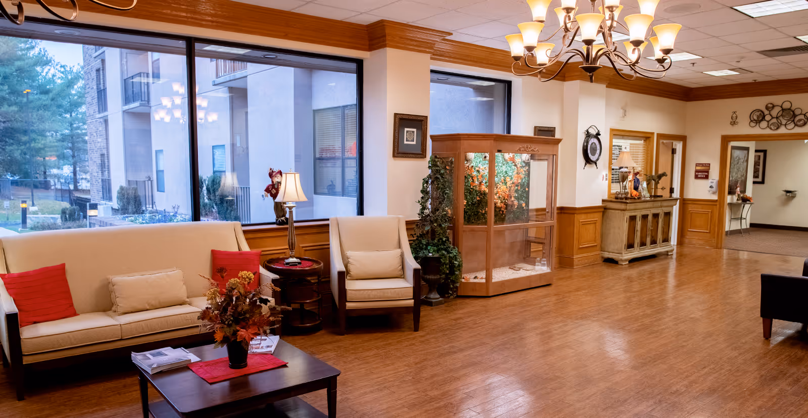 A spacious and well-lit living room area with large windows showing an outside view of a building and trees. The room features a beige sofa with red and beige cushions, a matching armchair, a wooden coffee table with a floral arrangement, a side table with a lamp, and a wooden display cabinet with plants inside. The floor is wooden, and a chandelier hangs from the ceiling. There is also a wooden sideboard with decorative items and a wall clock.