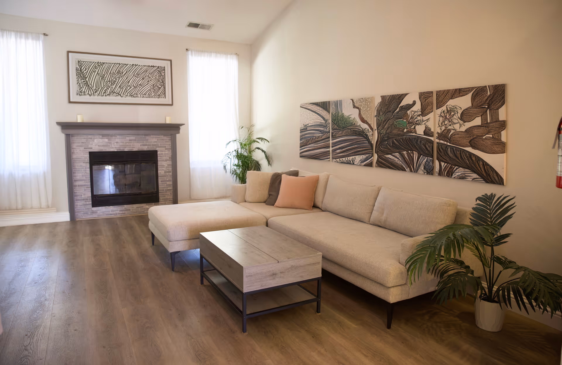 A cozy living room with a beige sectional sofa adorned with a peach and a gray pillow, a wooden coffee table with a metal frame, a fireplace with a gray mantel and tile surround, two tall windows with sheer white curtains, a potted plant near the sofa, and a three-panel botanical artwork on the wall.