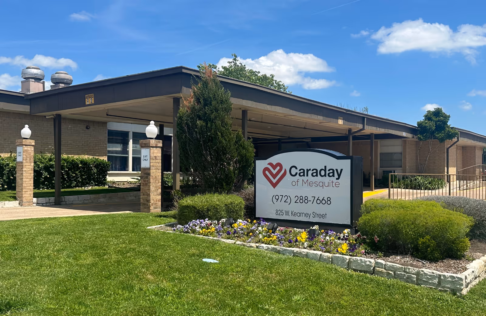 Exterior view of Caraday of Mesquite senior living facility with a covered entrance, green lawn, flower bed, and a sign displaying the facility name, phone number, and address under a blue sky with some clouds.