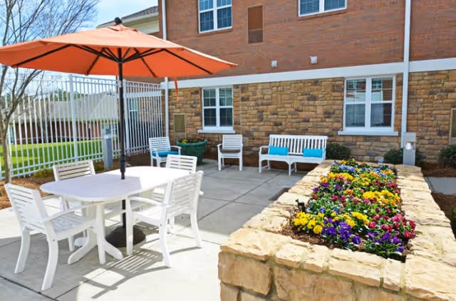 Outdoor patio area with white chairs and tables, a large orange umbrella providing shade, a stone planter filled with colorful flowers, and a brick building with windows in the background.