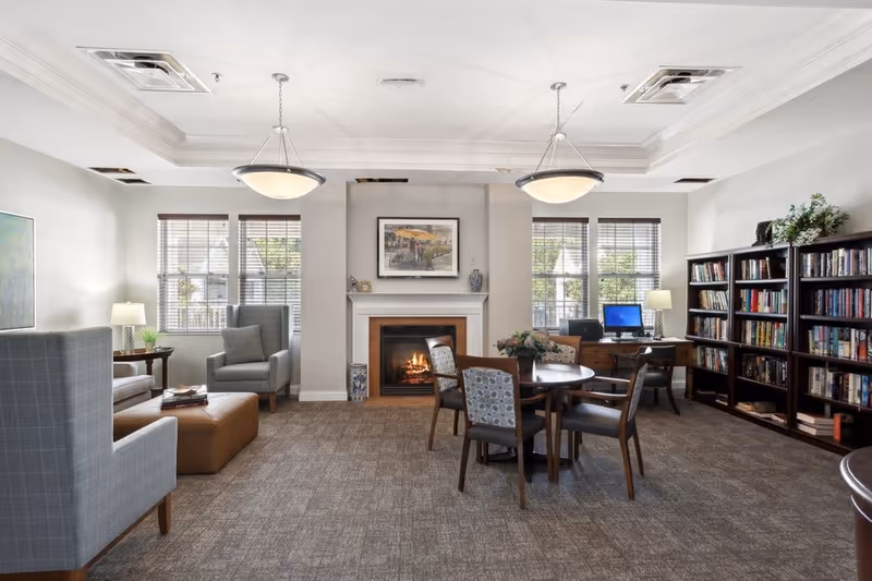 Bright communal living room with a fireplace, armchairs, a round table, desk with computer and bookshelves.