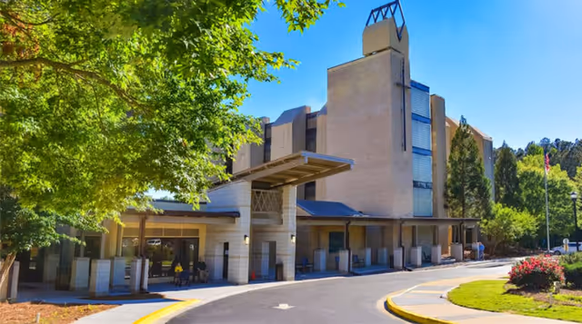 Exterior view of a multi-story assisted living facility building with a covered entrance, surrounded by trees and landscaping under a clear blue sky.
