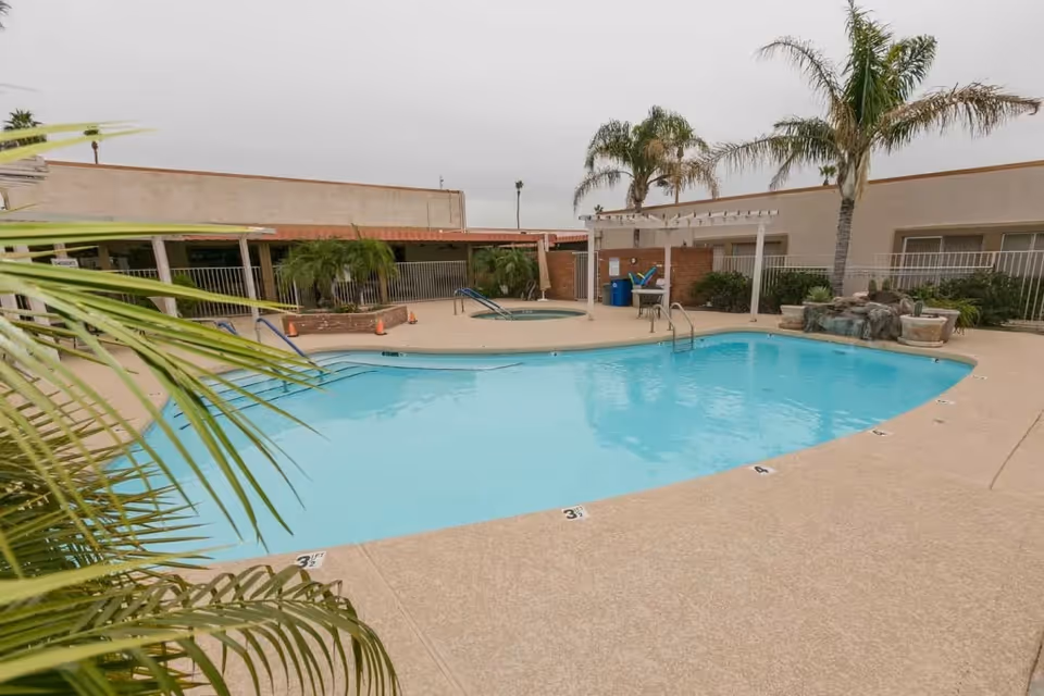 Outdoor swimming pool area at Olive Ridge Senior Living with clear blue water, surrounded by a concrete deck. There are palm trees and other greenery around the pool, a hot tub in the background, and a small waterfall feature on the right side. The area is enclosed by a fence and buildings.