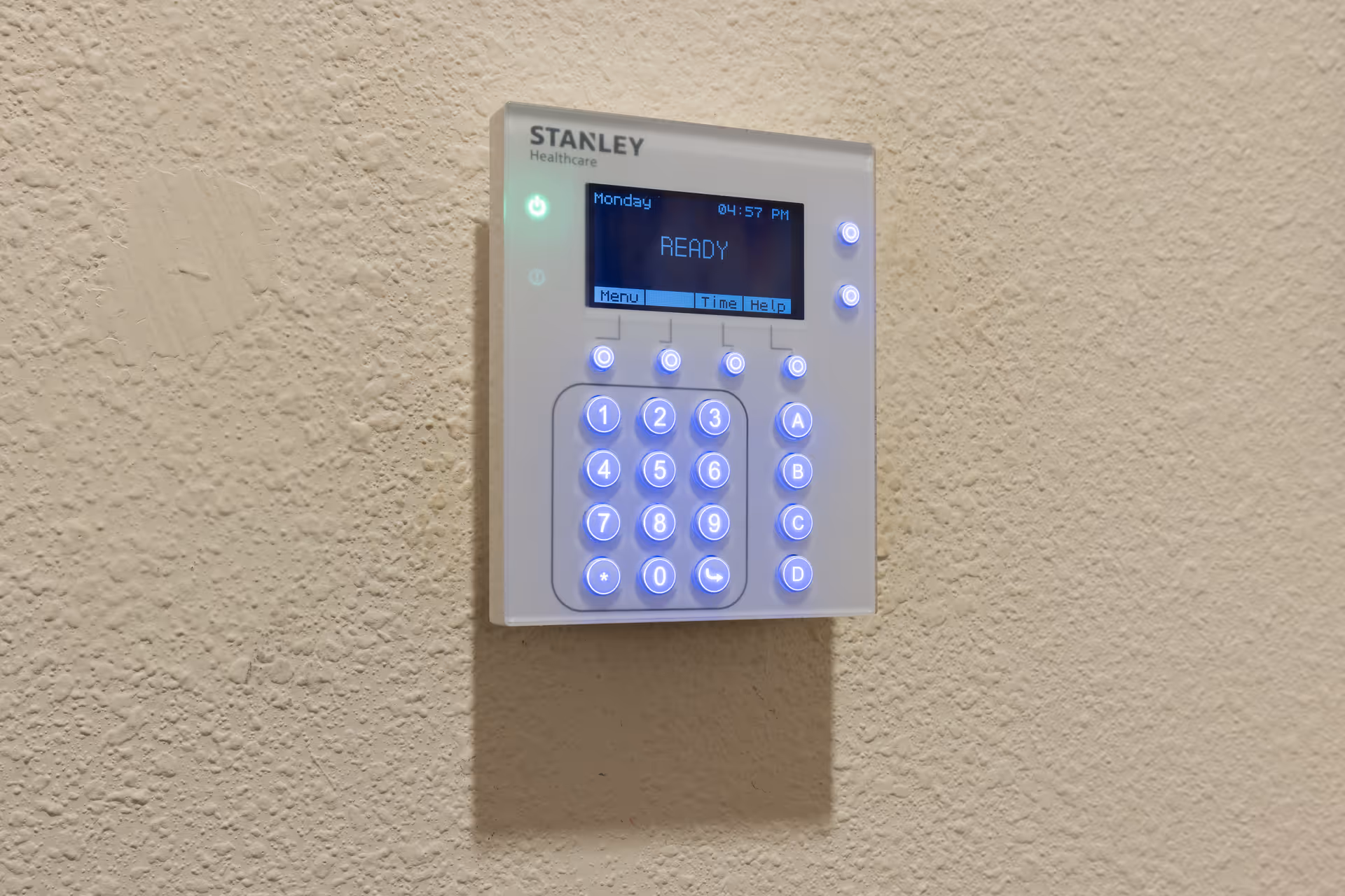 A Stanley Healthcare keypad mounted on a textured beige wall, displaying the word 'READY' on its blue screen with illuminated buttons.