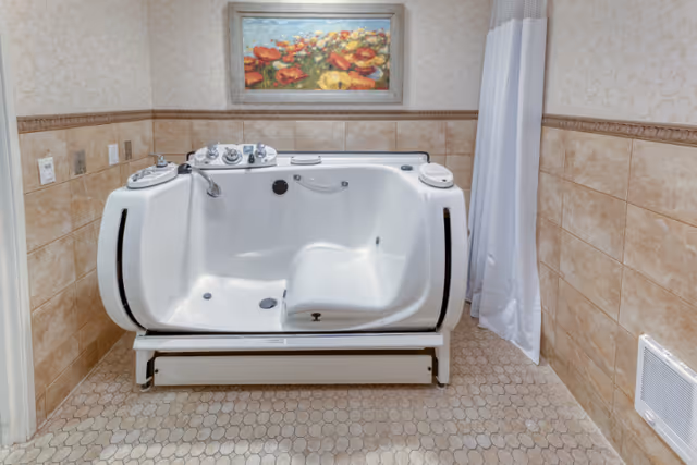 A walk-in bathtub with built-in seat and controls, situated in a tiled bathroom with beige hexagonal floor tiles and beige wall tiles. A floral painting hangs on the wall above the bathtub, and a white shower curtain is partially visible on the right side.