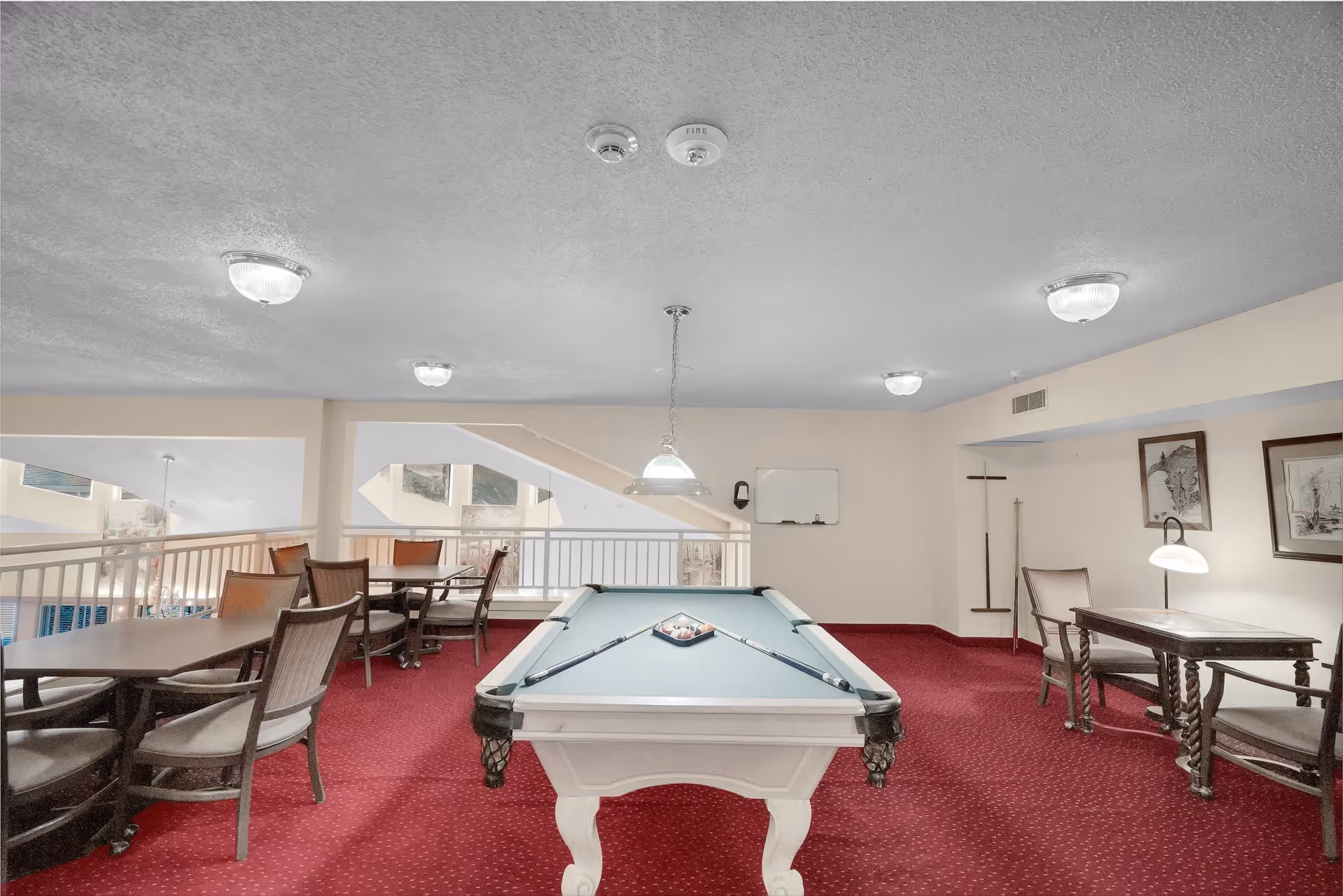 Interior room with a pool table in the center, surrounded by several tables and chairs. The room has a red carpet, beige walls, and ceiling lights. There are framed pictures on the wall and a whiteboard mounted on one wall.