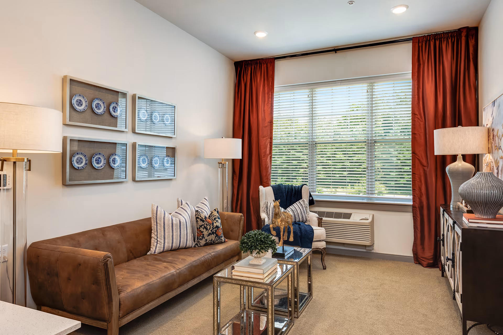 A cozy living room with a brown leather sofa adorned with striped and floral pillows, a white armchair with a blue throw blanket, two glass-top nesting tables with decorative items, two floor lamps, a window with red curtains and blinds, and framed decorative plates on the wall.