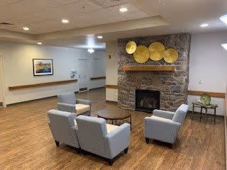 A cozy common area with four gray armchairs arranged around a small round wooden table in front of a stone fireplace. Above the fireplace are decorative golden circular wall hangings. The room has wood flooring, neutral-colored walls, and ceiling lights. A framed picture hangs on one wall, and a small table with a decorative item is placed near the fireplace.