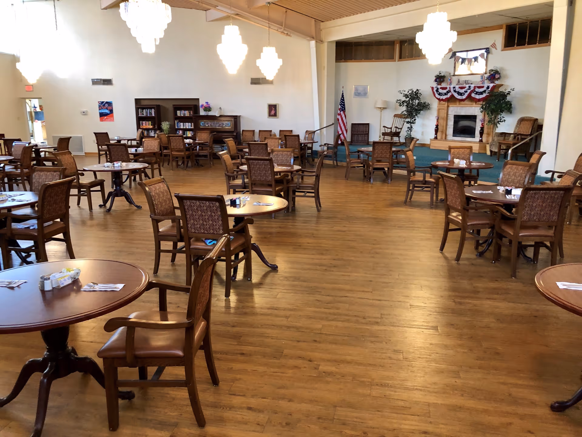 A spacious dining room with multiple round wooden tables and chairs arranged neatly on a wooden floor. The room features a fireplace decorated with patriotic bunting and flanked by plants, an American flag, bookshelves, and several chandeliers hanging from the ceiling.