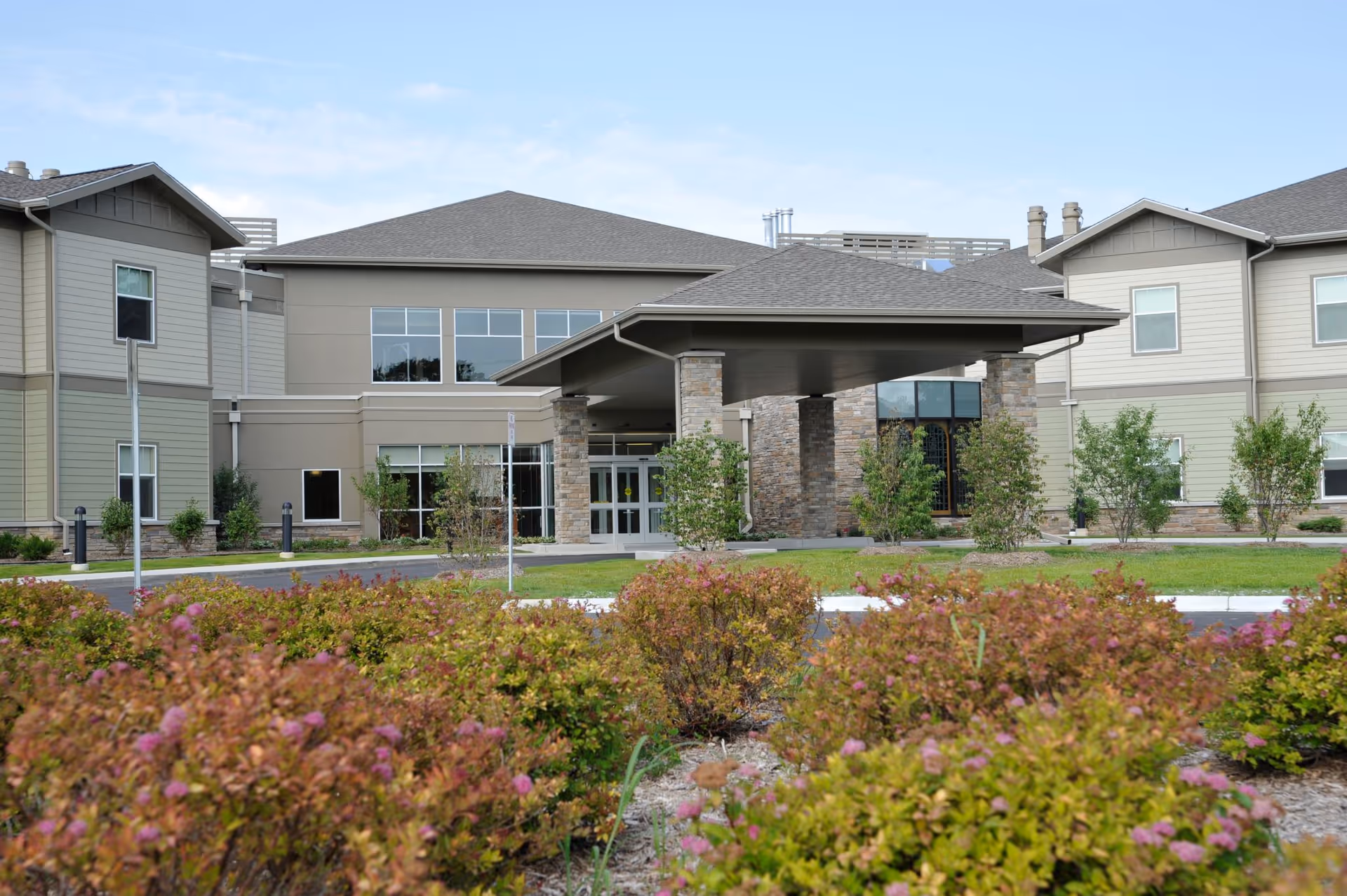 Exterior view of The Neighborhoods of White Lake senior living facility showing the main entrance with a covered drop-off area, surrounded by landscaped bushes and greenery under a partly cloudy sky.