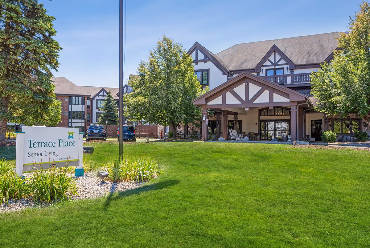 Exterior view of a senior living facility named Terrace Place with a well-maintained green lawn, trees, and a building featuring Tudor-style architecture under a clear blue sky.