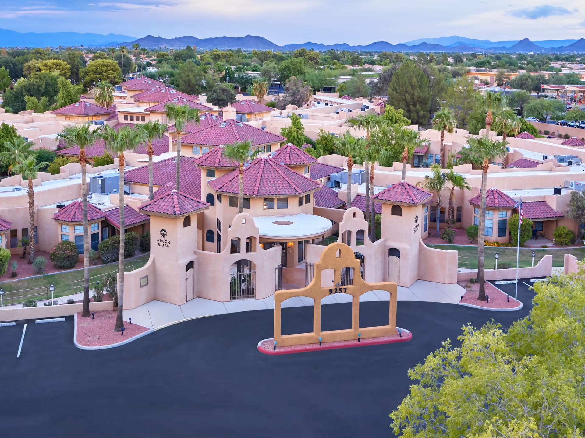 Aerial view of Arbor Ridge senior living facility featuring southwestern-style architecture with beige stucco walls and red tile roofs, surrounded by palm trees and greenery, with mountains visible in the background.