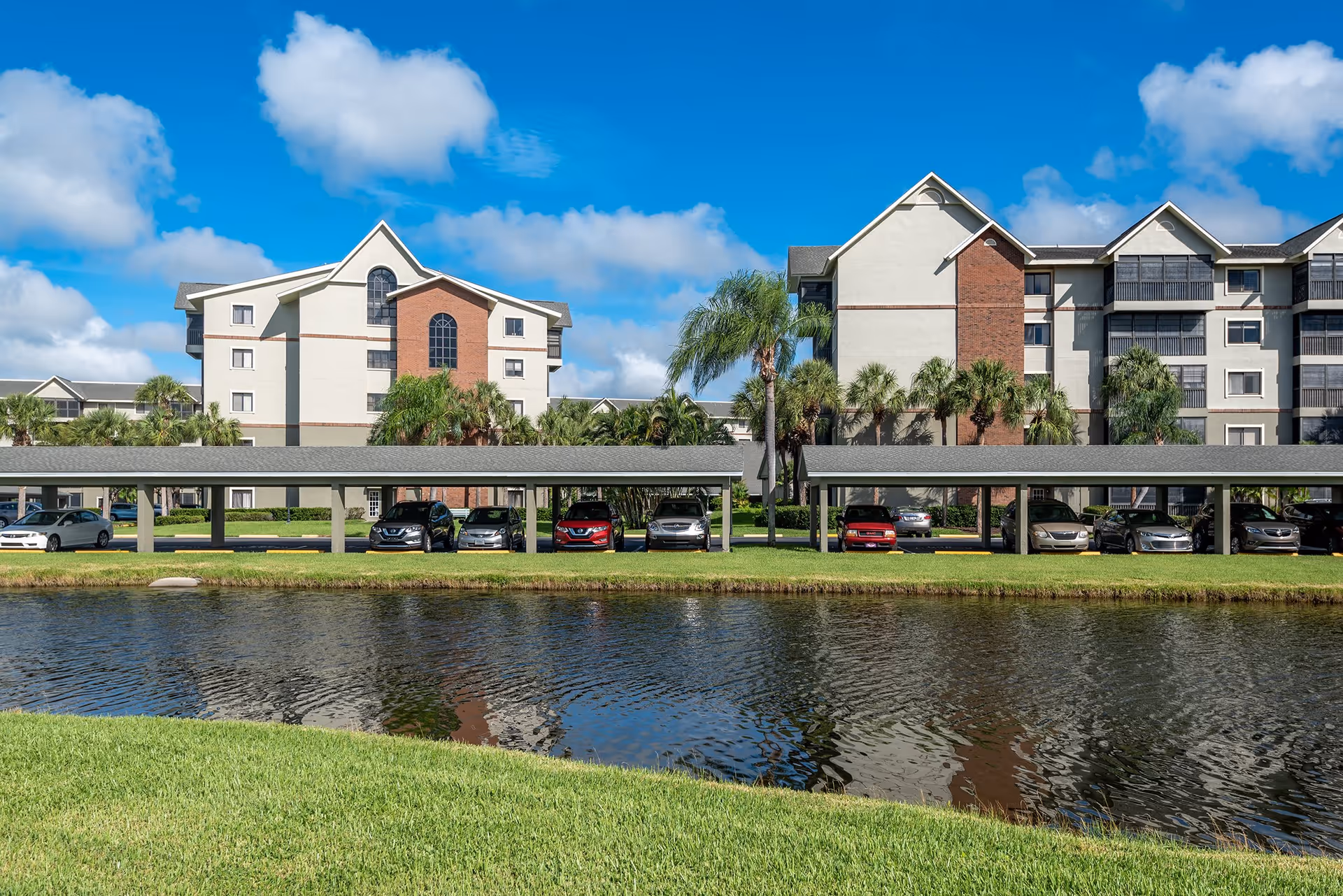 Exterior view of South Port Square senior living facility showing multi-story buildings with a mix of white and brick facades, covered parking spaces with cars, palm trees, and a pond in the foreground under a blue sky with scattered clouds.