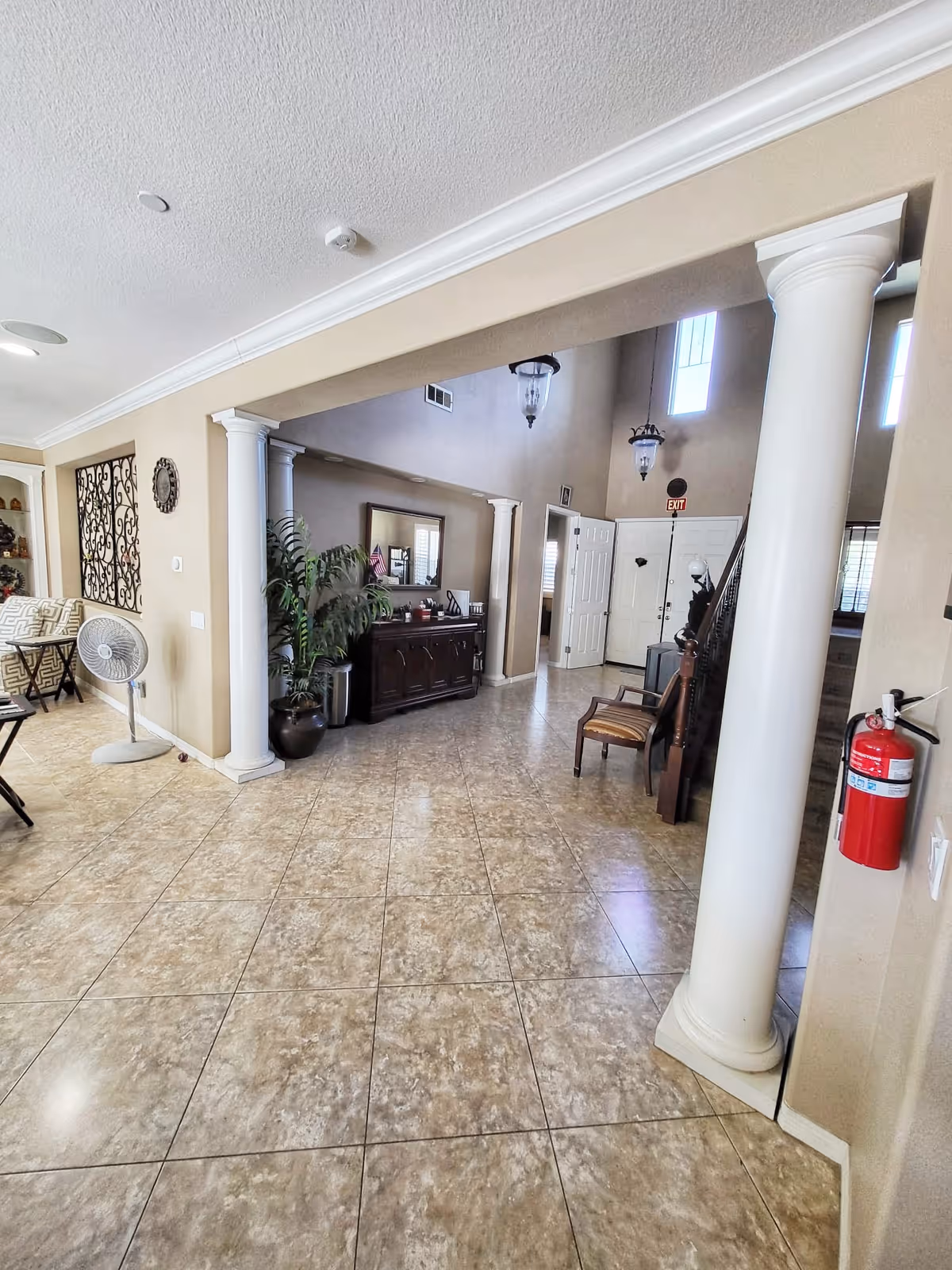 Interior view of a residential assisted living home featuring a spacious tiled floor area with white columns, a wooden sideboard with a mirror above it, a potted plant, a chair, and a staircase leading upstairs. The walls are painted beige, and there are hanging light fixtures and an exit sign above a white door.