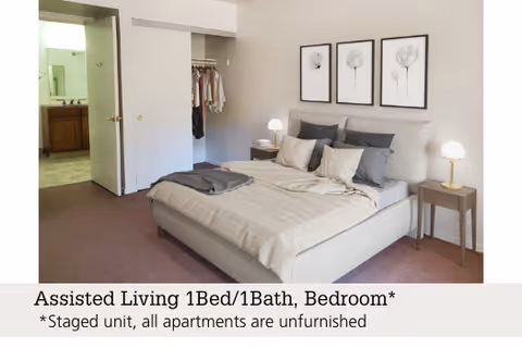 Staged one-bedroom with a made bed, nightstands and lamps, an open closet and a doorway leading to a bathroom.