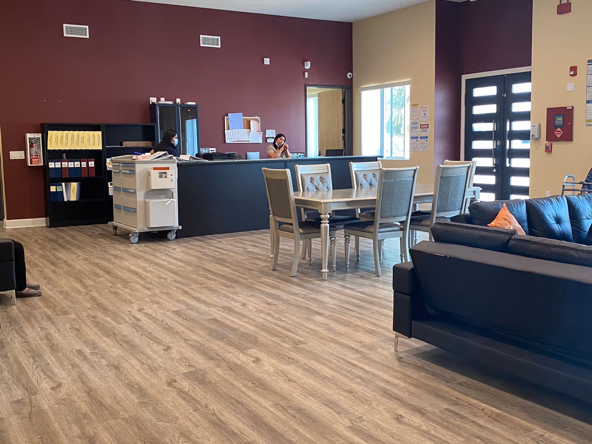 Interior view of a senior living facility reception area with a front desk where two staff members are working, one on the phone and the other wearing a mask. There is a dining table with six chairs in the middle of the room and a black leather couch with an orange pillow on the right side. The floor is wood-patterned, and the walls are painted in maroon and beige tones. A door with glass panels is visible in the background along with some office shelves and a fire extinguisher on the left wall.