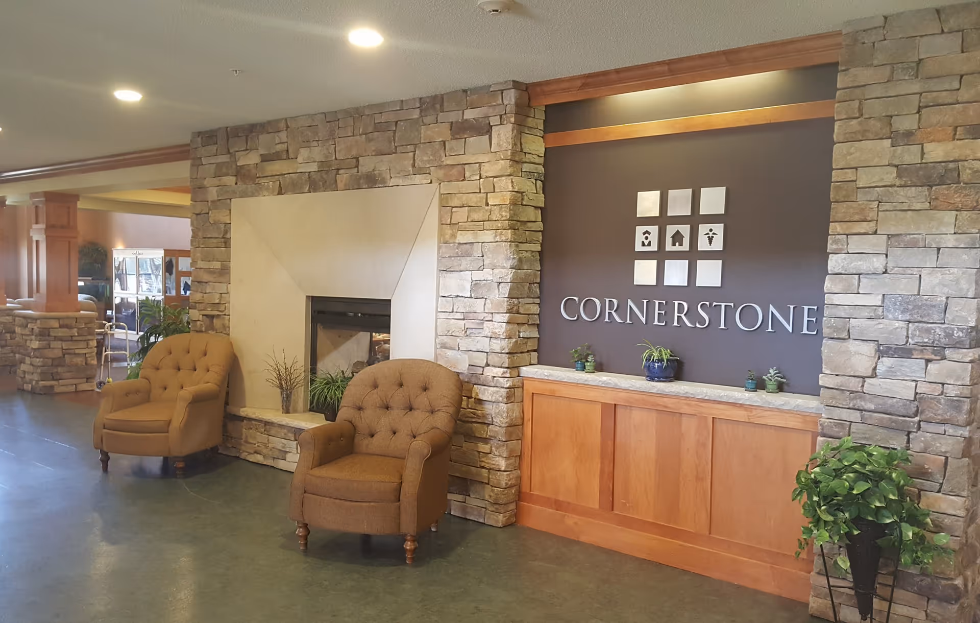 Interior view of a senior living facility lobby area with a stone fireplace, two brown upholstered armchairs, a wooden reception desk with small potted plants, and a wall sign that reads 'CORNERSTONE' with decorative icons above it.