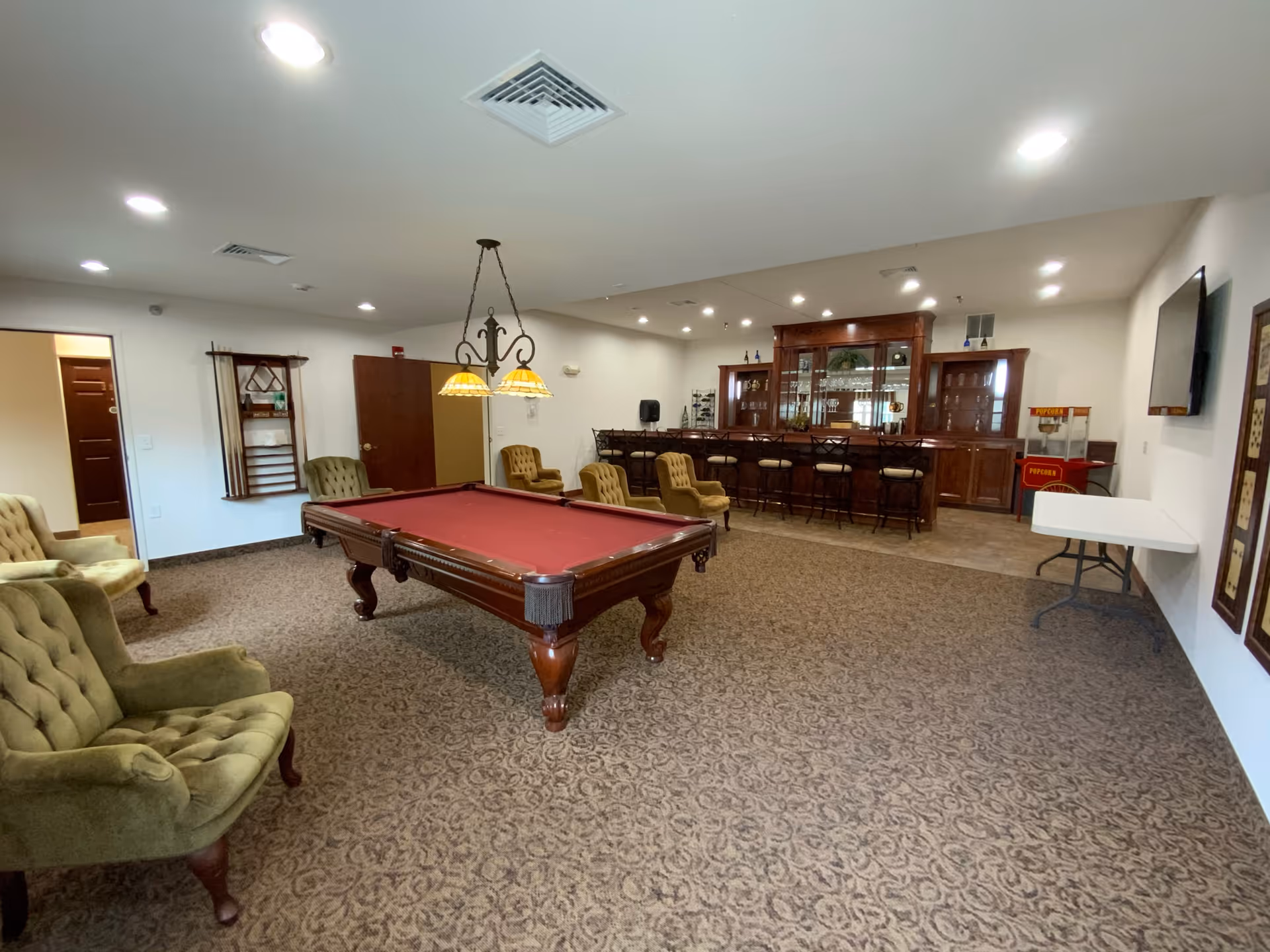 A spacious game room with a red felt pool table in the center, surrounded by several green upholstered armchairs. In the background, there is a wooden bar with bar stools and glass cabinets. The room has carpeted flooring, white walls, and ceiling lights. A popcorn machine and a folding table are visible on the right side, along with a wall-mounted TV.