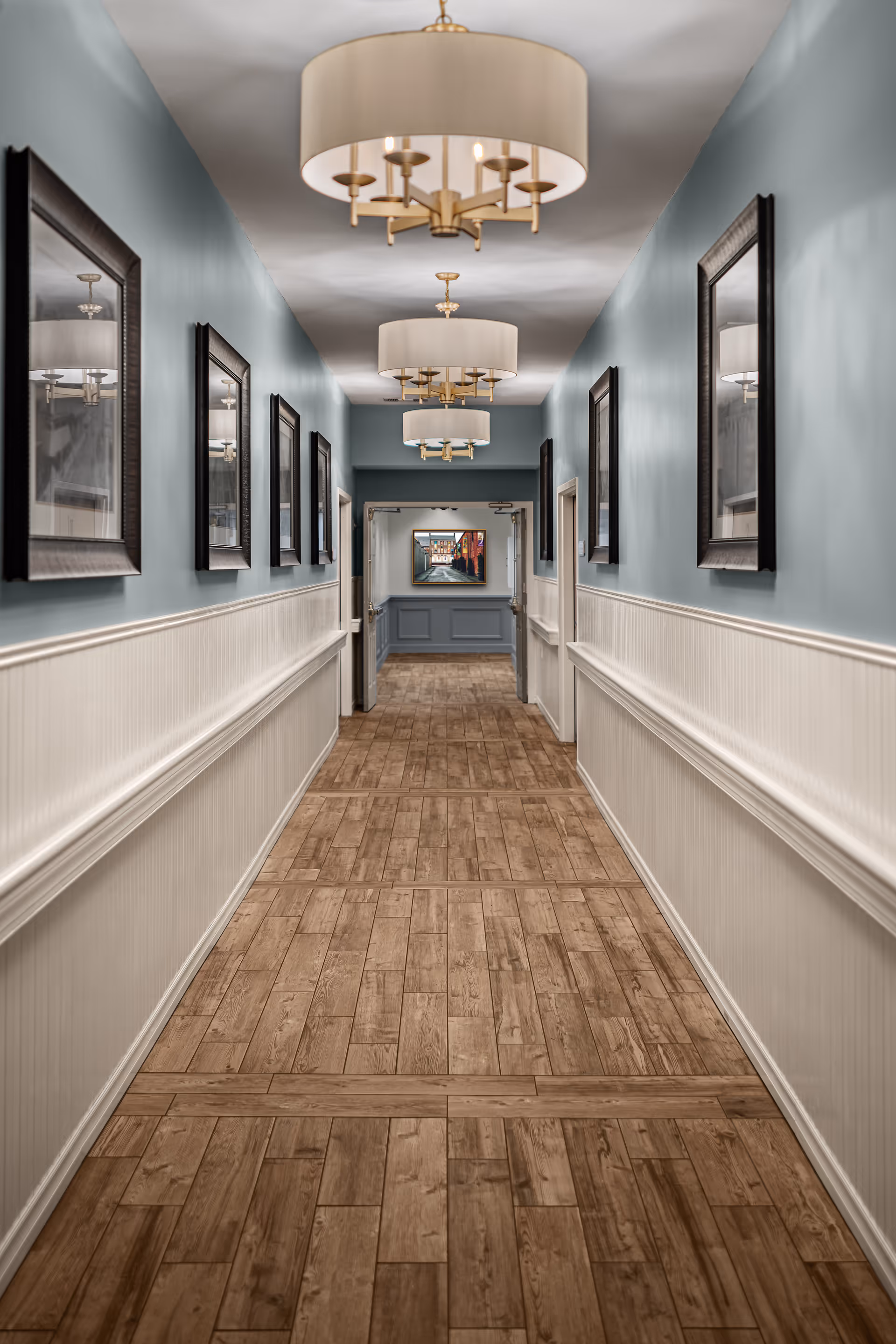 A long hallway with wooden flooring and light blue walls with white wainscoting. The hallway is decorated with framed pictures on both sides and has three large ceiling light fixtures with beige lampshades. At the end of the hallway, there is an open doorway leading to another room with a painting on the far wall.