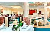 Elegant dining room with set tables, upholstered chairs, decorative columns and a white grand piano near a small stage.