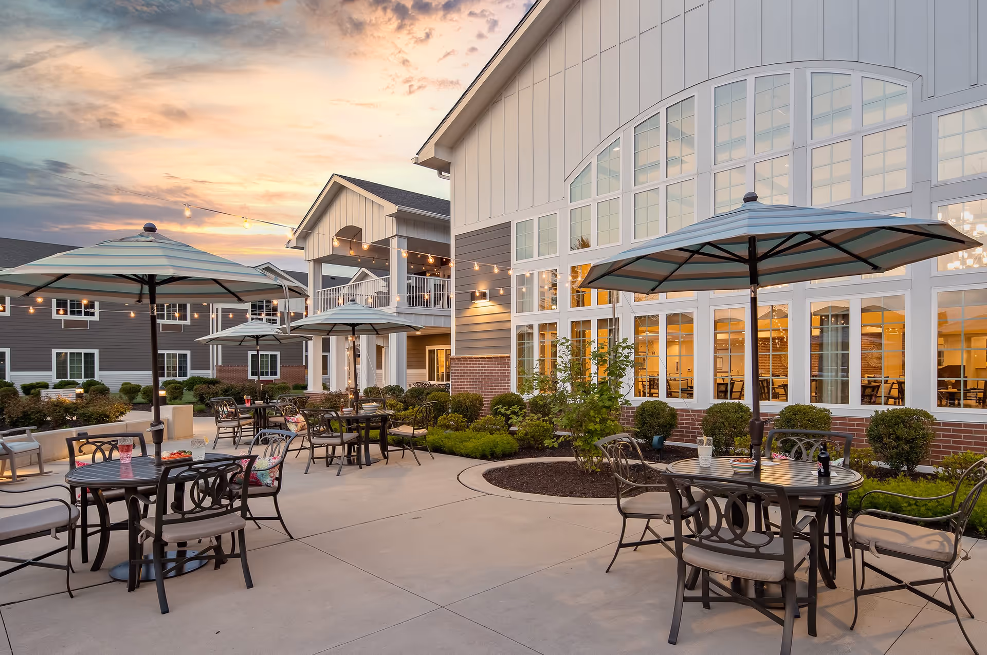 Outdoor patio area at The Province of Southampton with several round tables and chairs under large striped umbrellas. String lights are hung overhead, and the building with large windows is visible in the background during sunset.