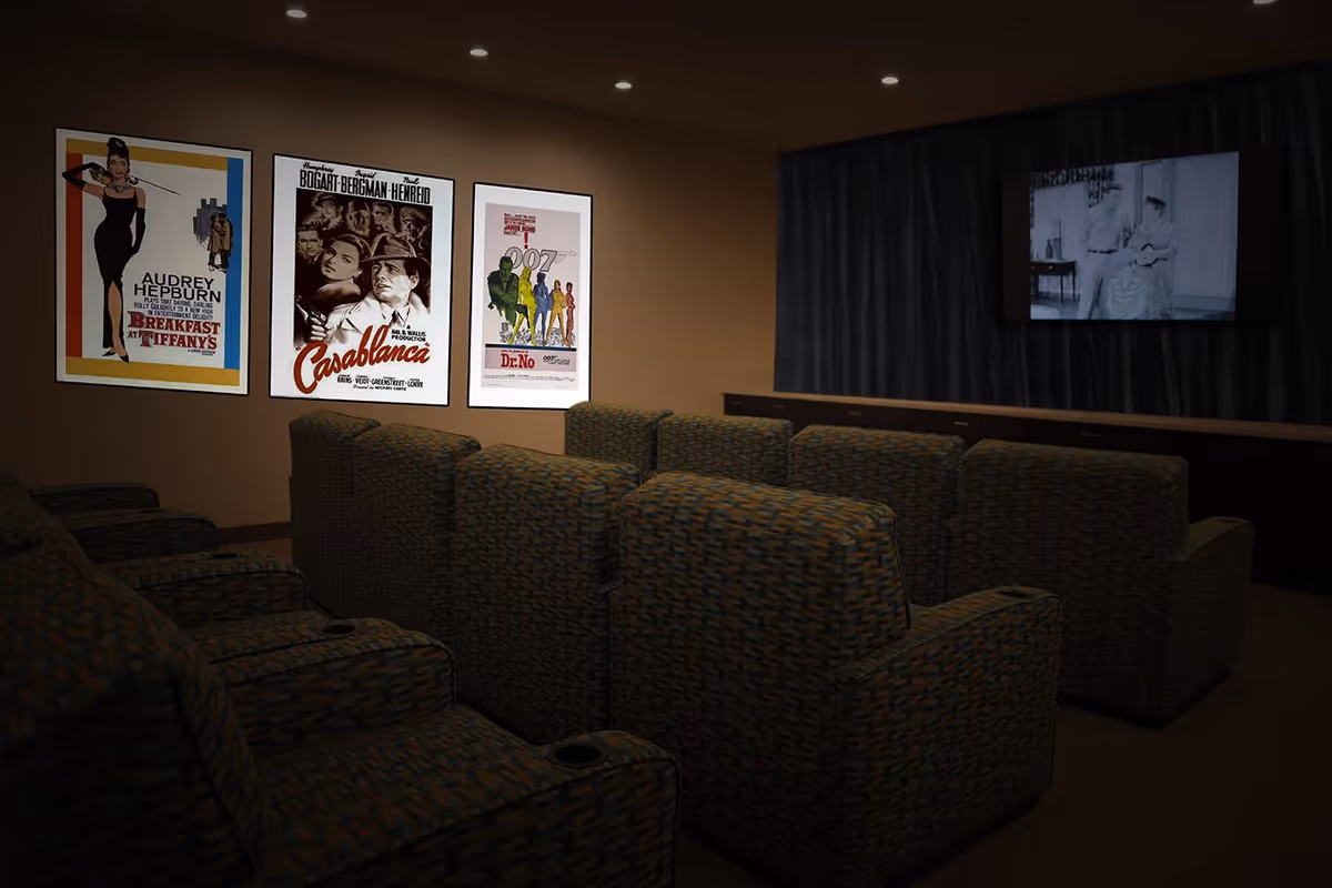 A cozy movie theater room with multiple rows of patterned cushioned seats facing a large screen showing a black and white film. The walls are decorated with framed classic movie posters including Breakfast at Tiffany's, Casablanca, and Dr. No.
