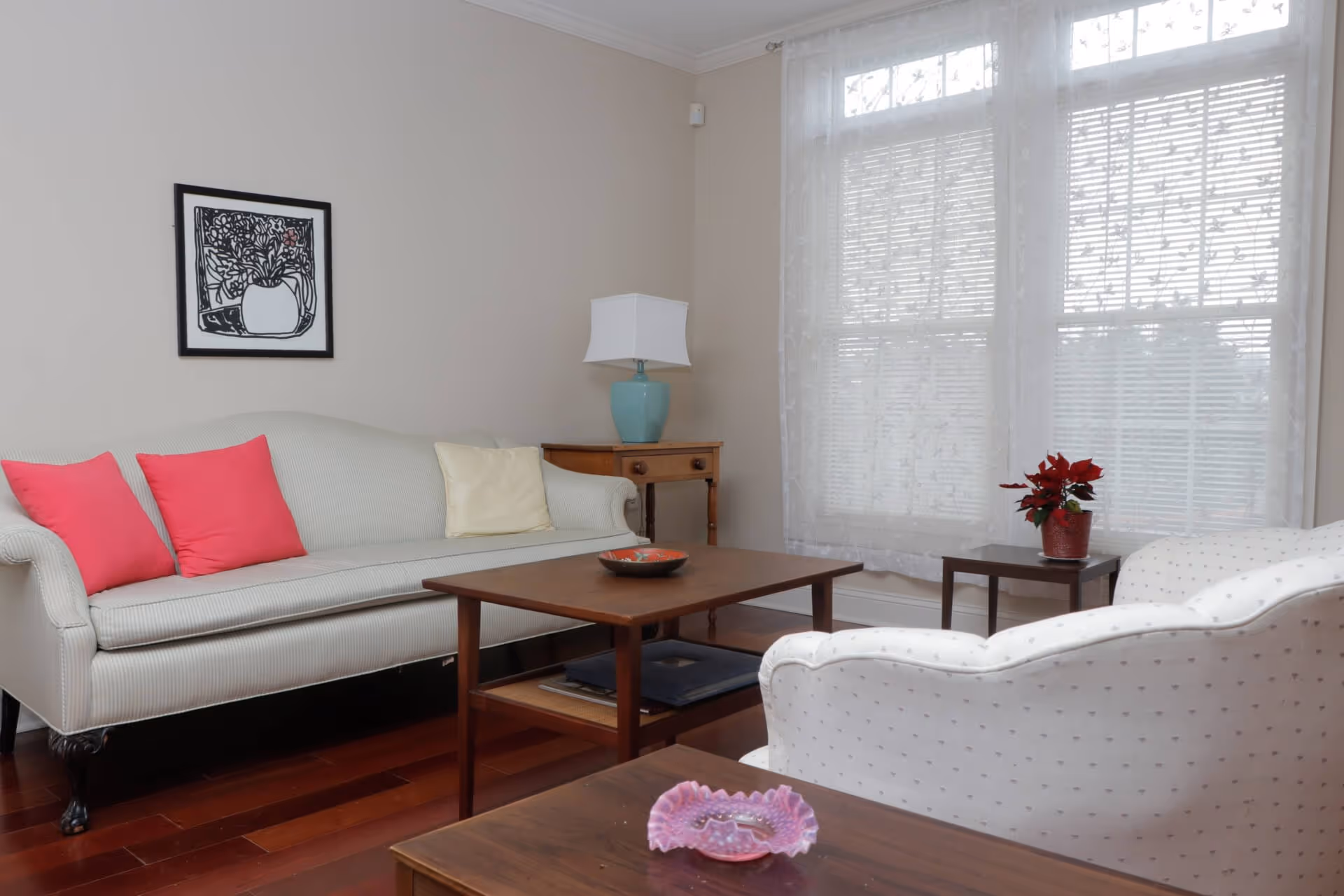 A cozy living room with a light-colored sofa adorned with two pink and one cream pillow, a wooden coffee table with a decorative bowl, a white armchair with subtle patterns, a small wooden side table with a red potted plant, a wooden console table with a blue lamp, and a large window with sheer white curtains allowing natural light to fill the room.