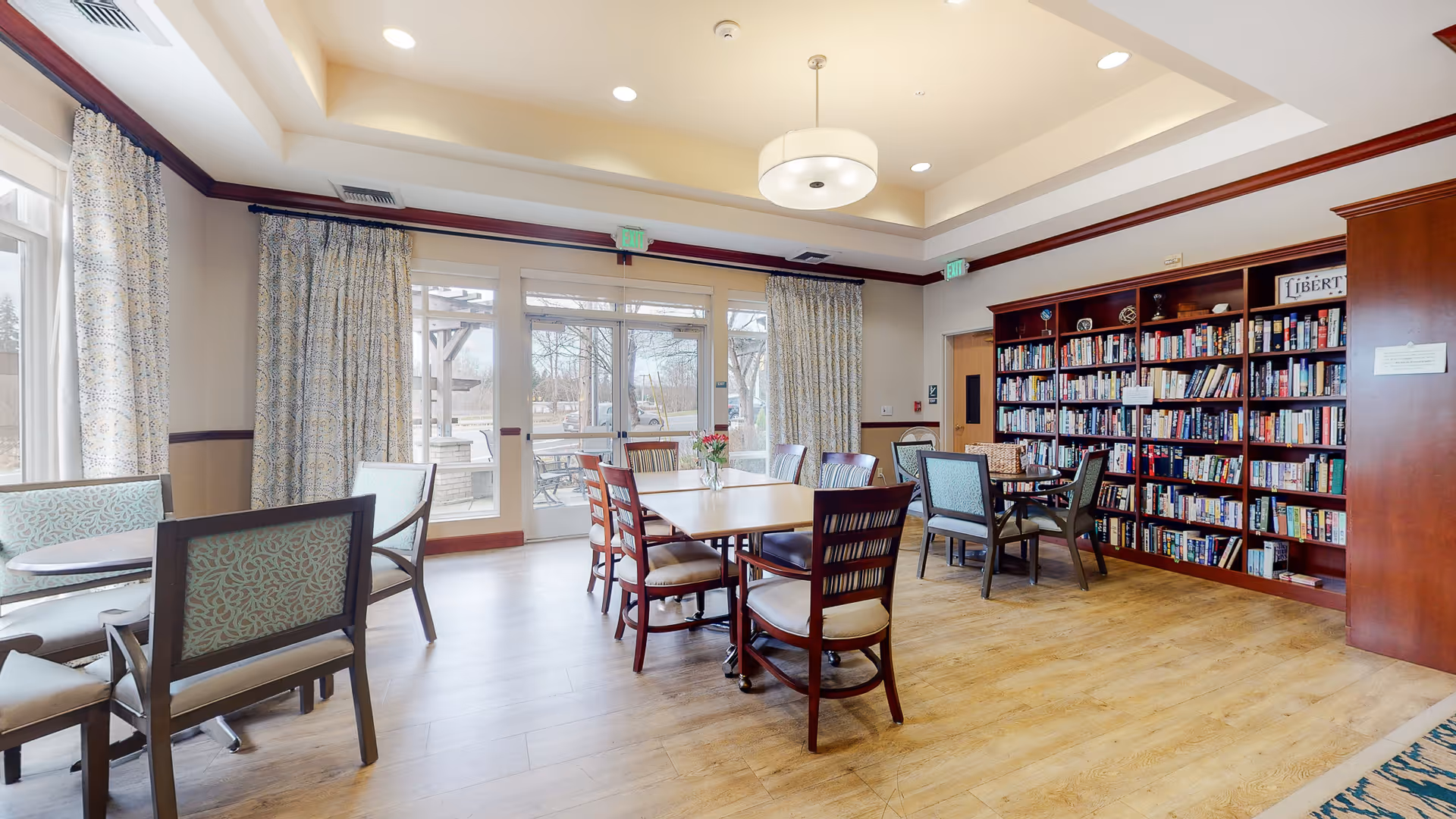 Sunlit communal room with dining tables and chairs and a large bookshelf against one wall.