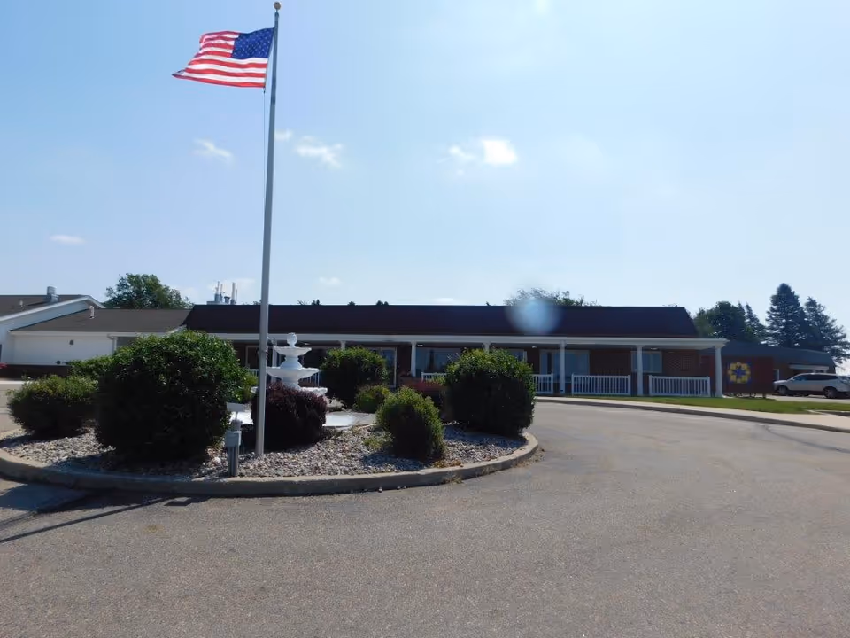 Front exterior of a single-story senior care building with an American flag, circular driveway, and landscaped fountain.