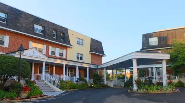 Exterior view of a senior living facility with a covered entrance driveway, brick and beige buildings, landscaped greenery, and a clear blue sky.